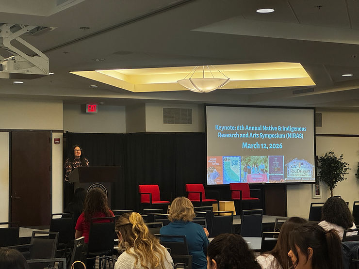 A presenter standing at a podium with a presentation screen to the back that reads, "Keynote: 6th Annual Native & Indigenous Research & Arts Symposium (NIRAS) and dated March 12, 2026