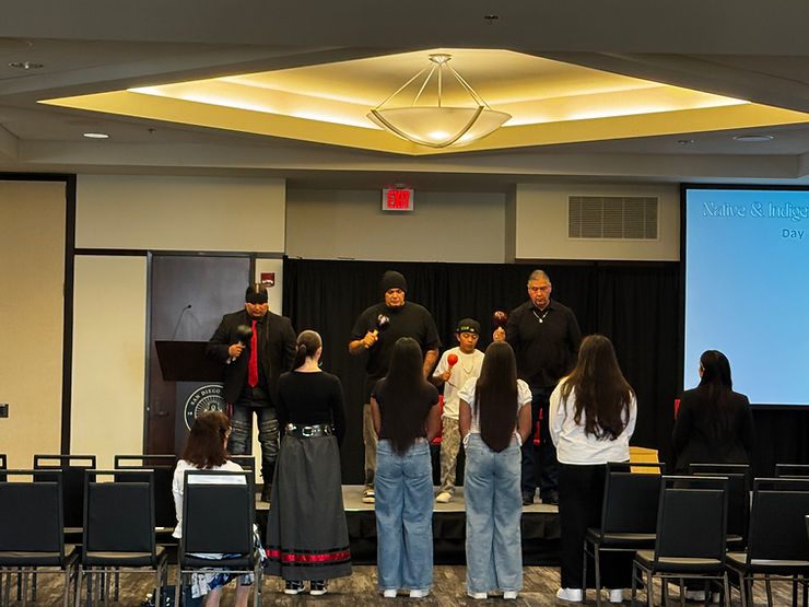 Photograph featuring individuals standing on a stage facing a row of individuals in front of them the floor.