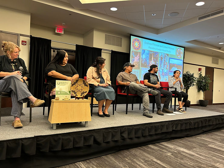 A photo of a panel discussion with six panelists seated on the stage with a presentation on display behind them